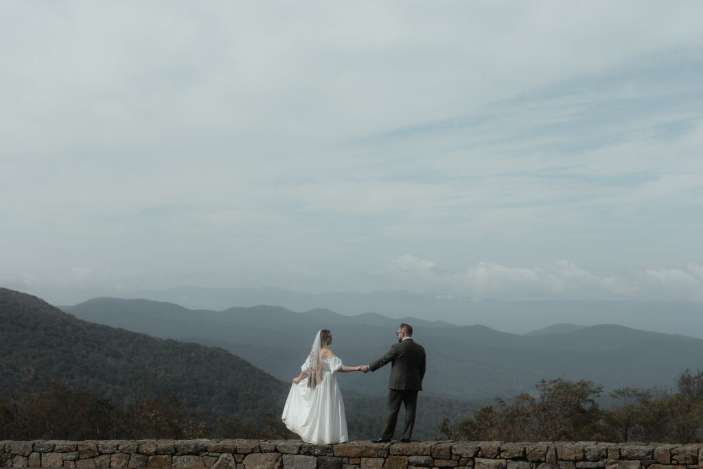 Eloping couple holding hands on a mountain ridge in Shenandoah National Park as wind moves through the bride’s dress beneath dramatic cloudy skies.