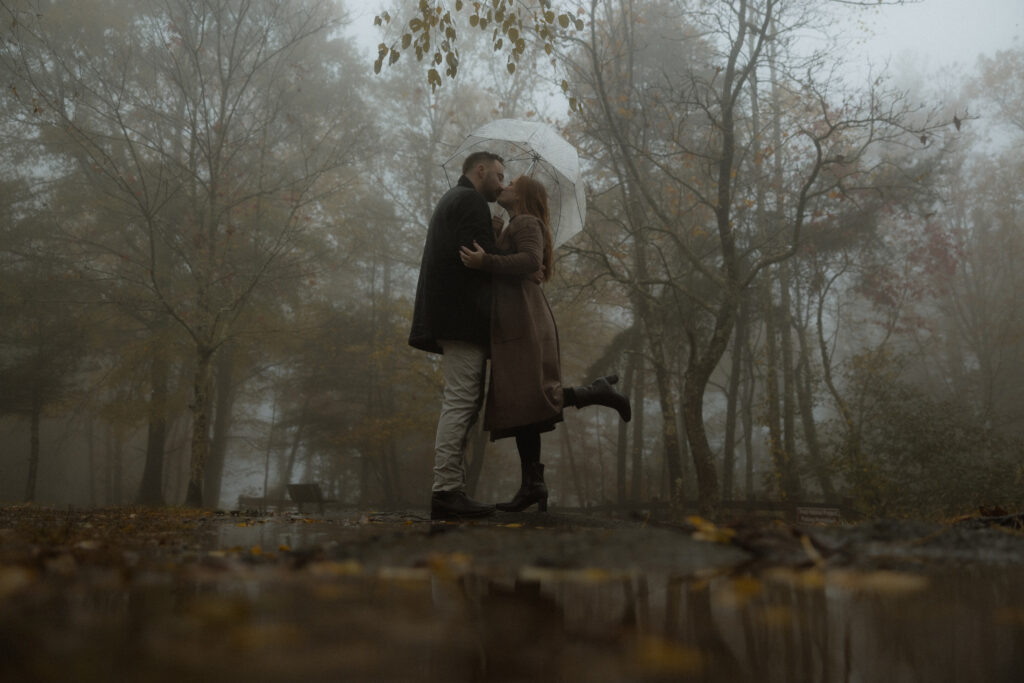 Couple walking hand in hand through fog with umbrellas during a rainy autumn engagement session at Amicalola Falls State Park in North Georgia