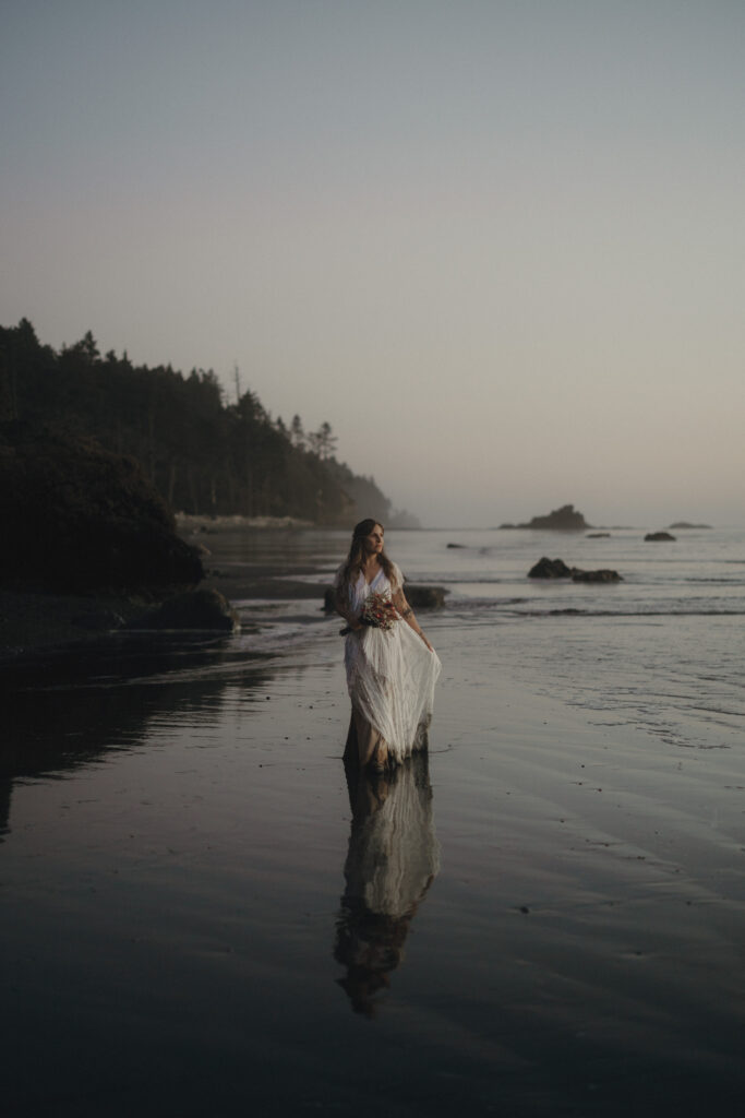 Bride’s dress blowing in ocean wind during a summer elopement on Ruby Beach in Olympic National Park with rugged coastline and sea stacks behind them.