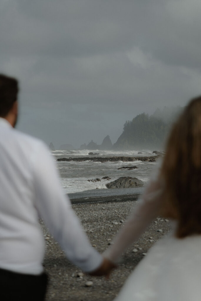 Eloping couple holding hands on Rialto Beach in Olympic National Park surrounded by coastal fog, driftwood, and rugged Pacific Northwest shoreline.
