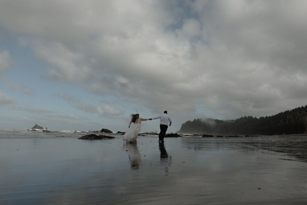 Eloping couple holding hands on Rialto Beach in Olympic National Park surrounded by coastal fog, driftwood, and rugged Pacific Northwest shoreline.