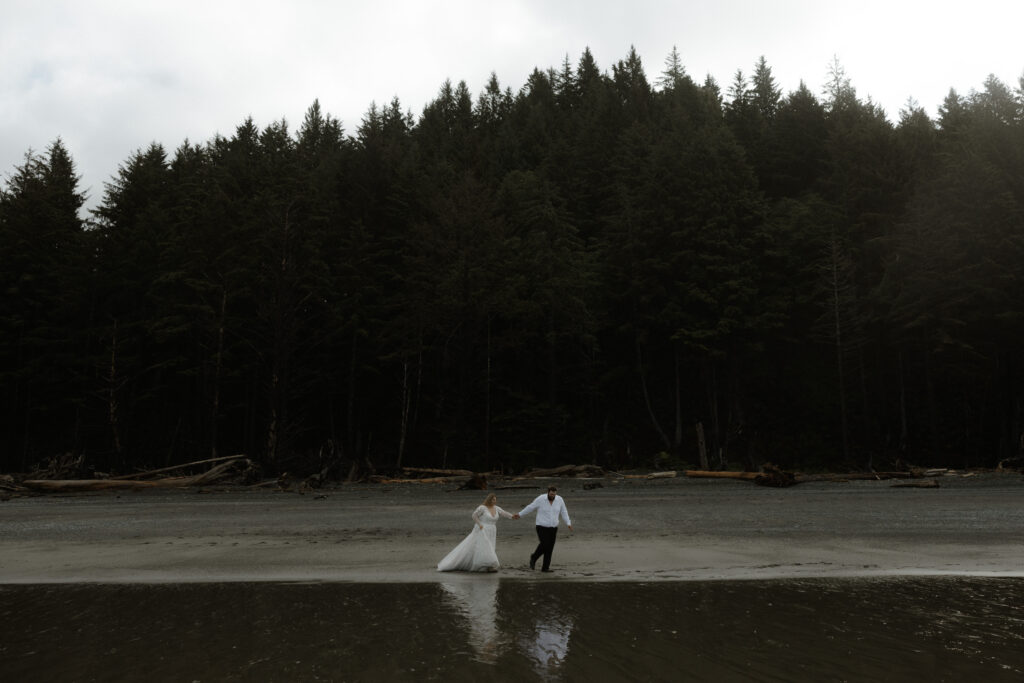 Eloping couple holding hands on Rialto Beach in Olympic National Park surrounded by coastal fog, driftwood, and rugged Pacific Northwest shoreline.