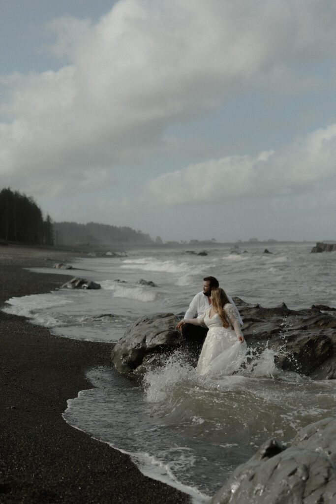 Eloping couple holding hands on Rialto Beach in Olympic National Park surrounded by coastal fog, driftwood, and rugged Pacific Northwest shoreline.