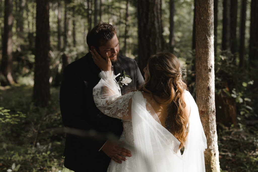 Bride and groom embracing along the shoreline of Lake Cushman with evergreen forests and mountain reflections in Olympic National Park, Washington.