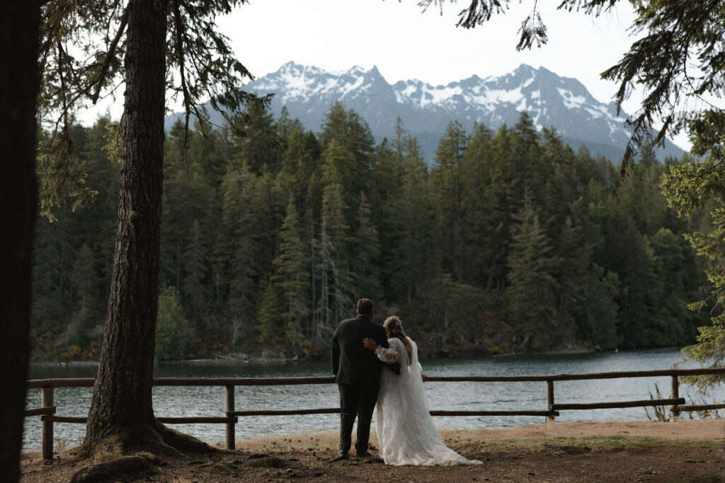 Bride and groom embracing along the shoreline of Lake Cushman with evergreen forests and mountain reflections in Olympic National Park, Washington.