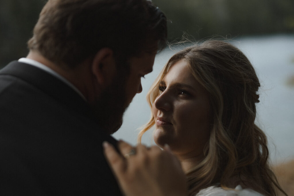 Bride and groom embracing along the shoreline of Lake Cushman with evergreen forests and mountain reflections in Olympic National Park, Washington.