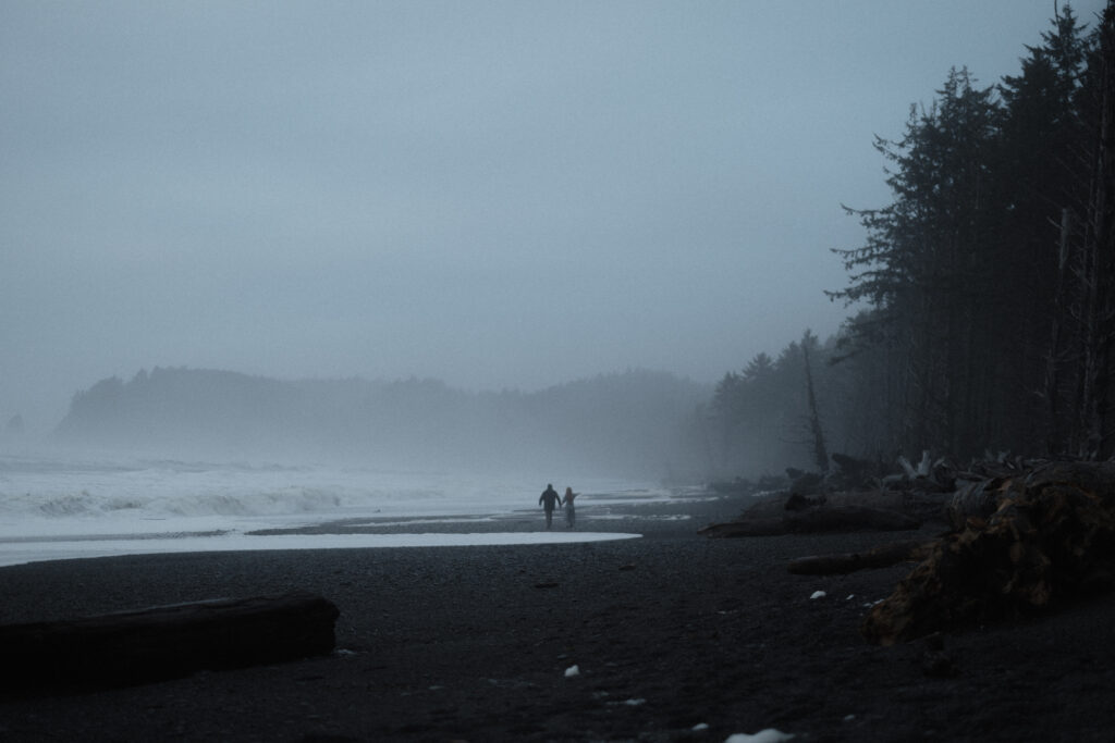 Bride and groom holding hands on Rialto Beach during a misty, overcast elopement with dramatic sea stacks fading into the fog.