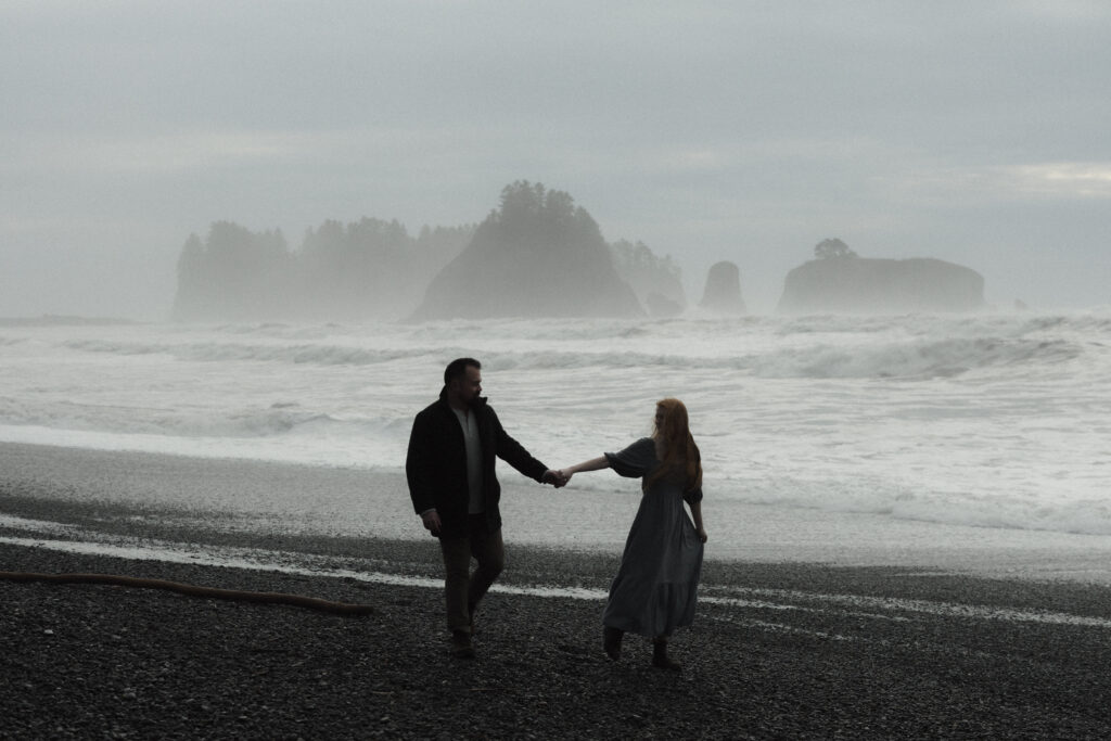 Bride and groom holding hands on Rialto Beach during a misty, overcast elopement with dramatic sea stacks fading into the fog.