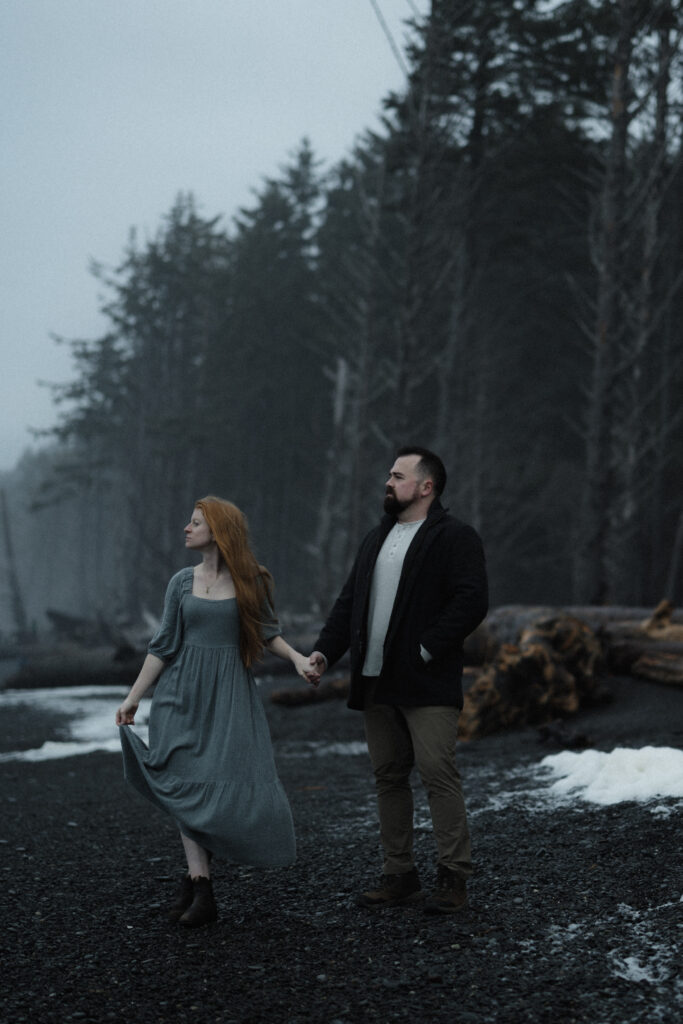 Bride and groom holding hands on Rialto Beach during a misty, overcast elopement with dramatic sea stacks fading into the fog.
